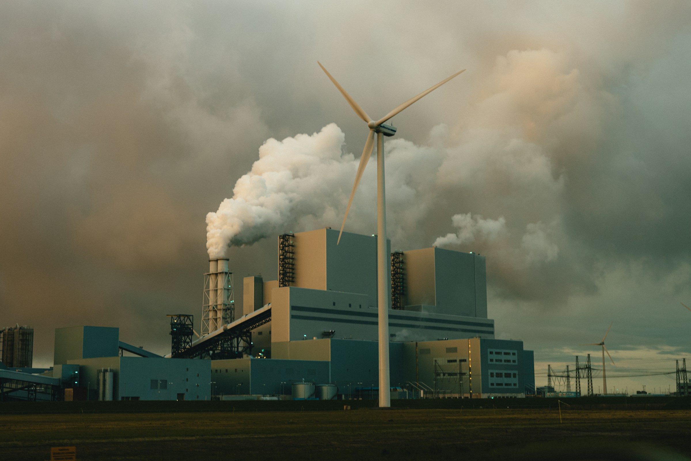 A tall wind turbine stands in the foreground in front of a boxy power plant with white smoke billowing from its chimneys.