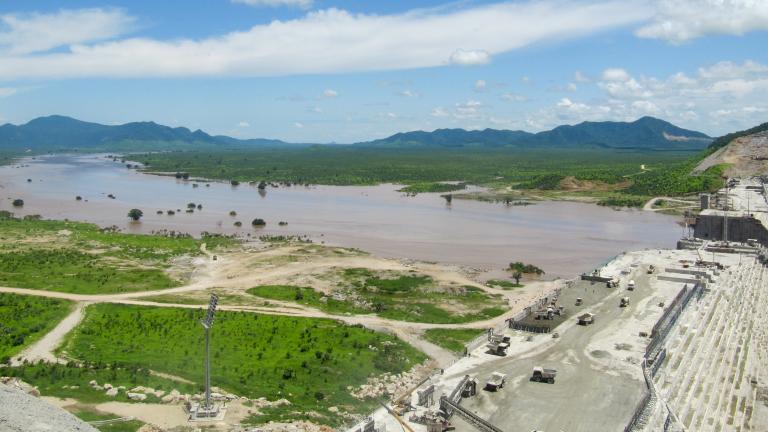 Photo of a large partially-completed dam in a broad river valley.