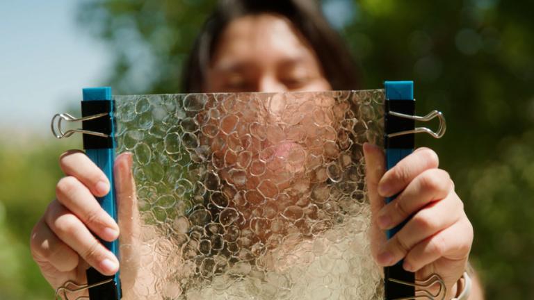 A person holds a translucent rectangle of material with scale-like shapes on it.