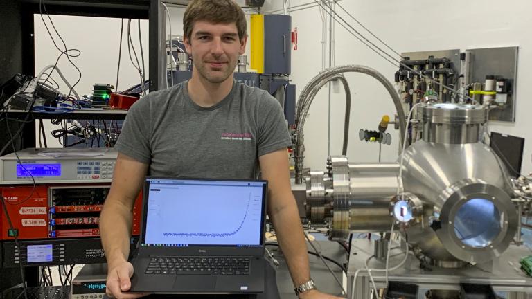 David Fischer sits beside the experiment’s vacuum chamber (illuminated in blue), where the high temperature superconductor tapes will be mounted for proton irradiation and in situ transport current measurement. His laptop shows data obtained in such measurements — the basis for determining the critical current.
