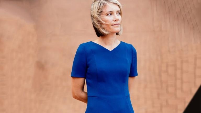 A person in a blue dress stands with a beige wall in the background