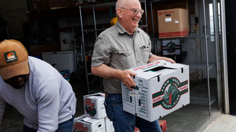 A man laughs while carrying a white and green box that reads "farm fresh vegetables."