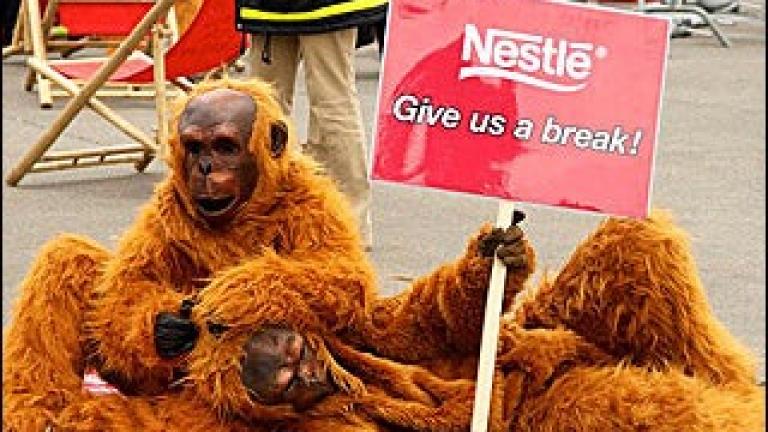 Two people in orangutan costumes on a sidewalk, one holding a sign reading "Nestle - Give us a break!"