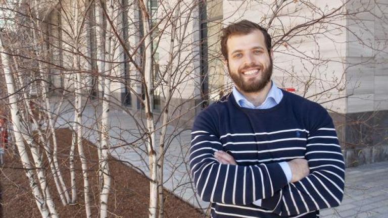 Photo of Theo Mouratidis, crossed arms, standing on MIT campus near birch trees and building