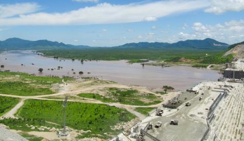 Photo of a large partially-completed dam in a broad river valley.