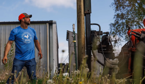 A person in a blue shirt and a baseball cap looks at a tractor that's installing fenceposts in a field.