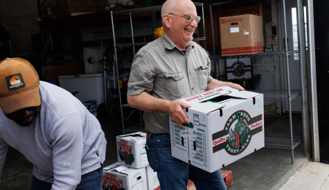 A man laughs while carrying a white and green box that reads "farm fresh vegetables."