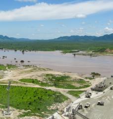 Photo of a large partially-completed dam in a broad river valley.
