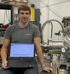 David Fischer sits beside the experiment’s vacuum chamber (illuminated in blue), where the high temperature superconductor tapes will be mounted for proton irradiation and in situ transport current measurement. His laptop shows data obtained in such measurements — the basis for determining the critical current.