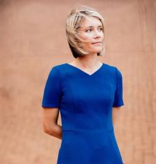 A person in a blue dress stands with a beige wall in the background