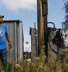 A person in a blue shirt and a baseball cap looks at a tractor that's installing fenceposts in a field.