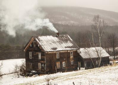 A dark wood clad house with snow visible on a standing-seam metal roof. Smoke is coming from the chimney. Snow-covered fields and winter forest are in the background.