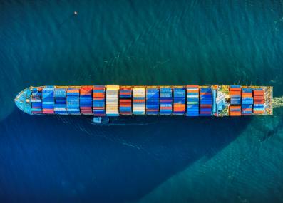 A large vessel full of shipping containers, viewed from above, crosses the water.